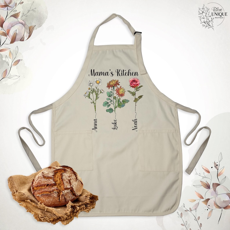 May include: Beige apron with the text "Mama's Kitchen" and floral illustrations. The apron features three flowers with the names "Anna", "Luke", and "Noah" below them. It has two pockets and a neck strap. A loaf of bread rests on a burlap cloth.