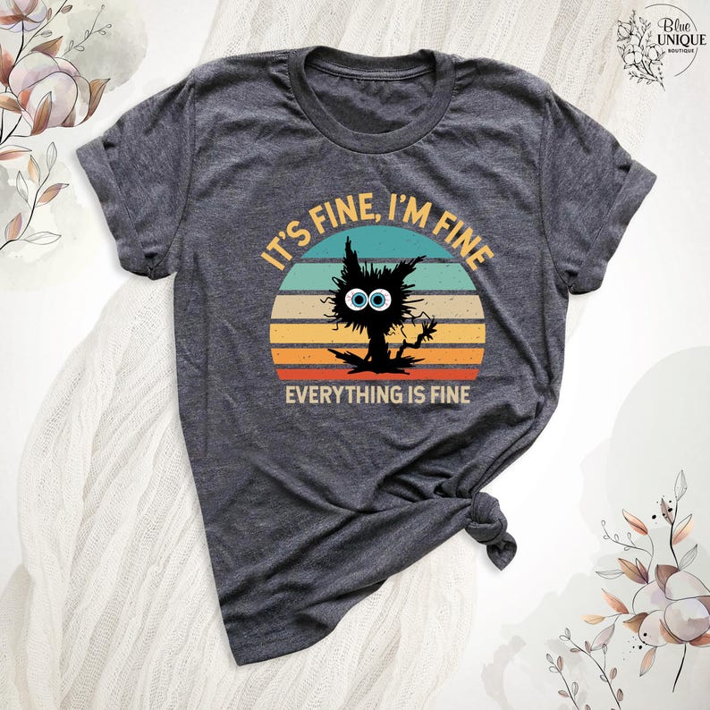 May include: A dark gray t-shirt with a vintage-style sunset graphic featuring a black cat with large eyes. The text "It's fine, I'm fine, everything is fine" is printed on the shirt.