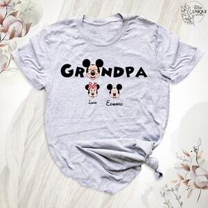 Puede incluir: Camiseta gris con la palabra "Grandpa" en letras negras. Mickey Mouse aparece en la palabra "Grandpa" con Minnie Mouse y otro Mickey Mouse debajo de la palabra. Los nombres "Luna" y "Edward" est&aacute;n escritos debajo de los personajes.