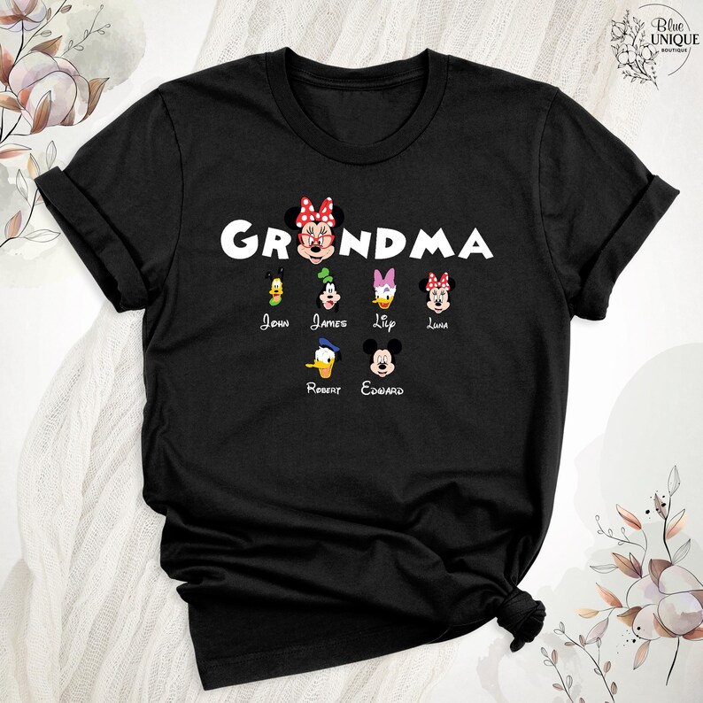 Puede incluir: Camiseta negra con la palabra "Grandma" en letras blancas y un lazo rojo. Hay seis personajes de dibujos animados debajo de la palabra, cada uno con un nombre debajo: John, James, Lily, Luna, Robert y Edward.