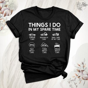 Op de afbeelding: Zwart T-shirt met witte tekst die "Things I do in my spare time" luidt. De tekst wordt omringd door zes witte pictogrammen die auto's voorstellen, waaronder rijden, onderzoek doen, zorgen voor, praten over, nadenken over en dromen over auto's.