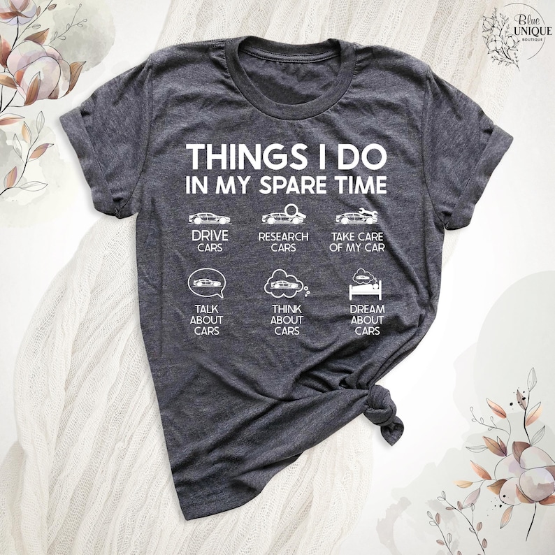 Op de afbeelding: Een donkergrijs T-shirt met een witte afbeelding waarop staat "Things I do in my spare time" en activiteiten worden opgesomd die te maken hebben met auto's. De activiteiten omvatten autorijden, onderzoek doen, zorgen voor, praten over, nadenken over en dromen over auto's.