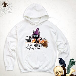 May include: A white hooded sweatshirt featuring a graphic of a black creature wearing a purple witch hat, riding a broomstick surrounded by flames. The text reads: IT IS FINE I AM FINE Everything is fine. The hoodie has long sleeves and a front pocket.
