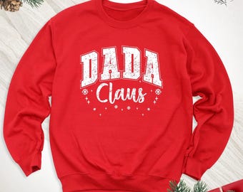 May include: A bright red crewneck sweatshirt featuring the white distressed text DADA Claus across the chest. The design includes small white star and snowflake accents surrounding the lettering, creating a festive holiday appearance.