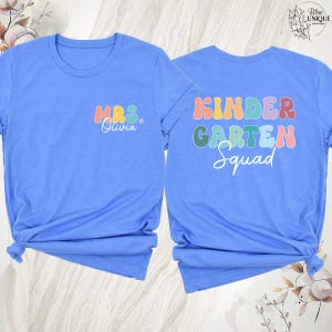 May include: Two light blue t-shirts. One shirt has "MRS. Olivia" in colorful text on the front. The other shirt has "KINDERGARTEN Squad" in colorful text on the back. Both shirts are tied at the bottom.