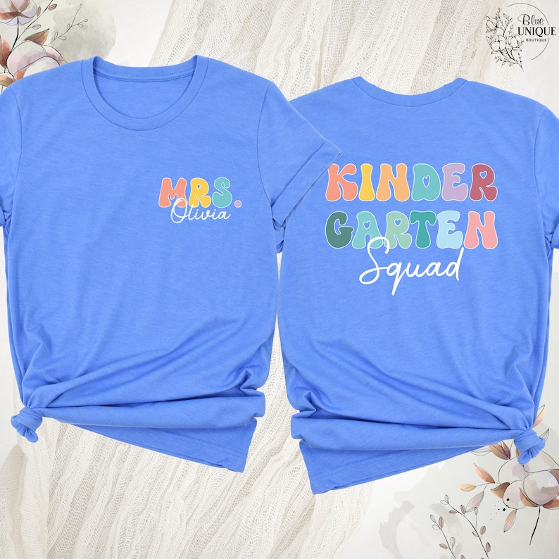 May include: Two light blue t-shirts. One shirt has "MRS. Olivia" in colorful text on the front. The other shirt has "KINDERGARTEN Squad" in colorful text on the back. Both shirts are tied at the bottom.