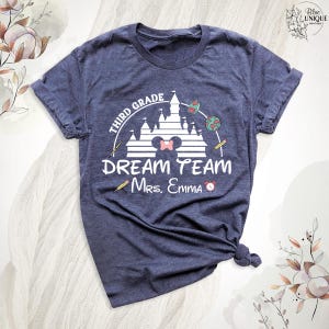 May include: Navy blue t-shirt with a white graphic featuring a castle, a Minnie Mouse bow, and the words "Third Grade Dream Team Mrs. Emma." Includes pencils and a clock graphic. A teacher-themed apparel item.