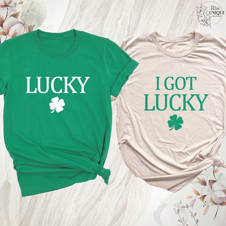 K&ouml;nnte beinhalten: Zwei T-Shirts, eines gr&uuml;n und eines beige, mit St. Patrick's Day-Motiven. Das gr&uuml;ne Shirt hat das Wort "LUCKY" in Wei&szlig; mit einem wei&szlig;en Kleeblatt. Das beige Shirt sagt "I GOT LUCKY" in Gr&uuml;n mit einem gr&uuml;nen Kleeblatt. Beide Shirts sind kurz&auml;rmelig.