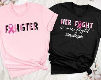May include: A light pink t-shirt with the word FIGHTER in black letters, featuring a pink ribbon as the letter I. A black t-shirt with the text HER FIGHT is our fight and #TeamSophia in white, with a pink ribbon replacing the letter I.