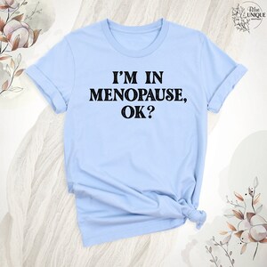May include: A light blue t-shirt with the text "I'm in Menopause, OK?" printed in black.