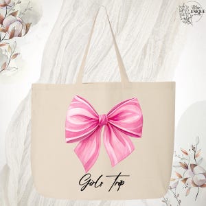May include: A beige tote bag featuring a large pink bow illustration with white accents. The words "Girls Trip" are written in a script font below the bow. The bag has two handles and is made of a canvas-like material.