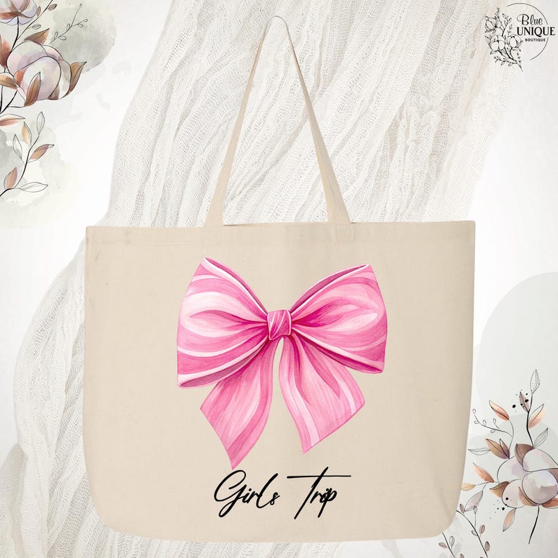 May include: A beige tote bag featuring a large pink bow illustration with white accents. The words "Girls Trip" are written in a script font below the bow. The bag has two handles and is made of a canvas-like material.