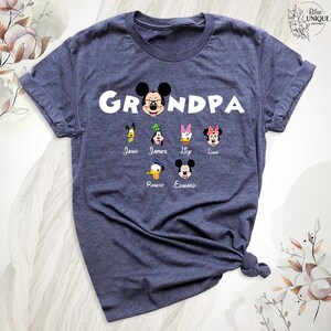 Puede incluir: Una camiseta azul marino con la palabra "Grandpa" en letras blancas. La camiseta presenta una imagen de dibujos animados de Mickey Mouse con otros seis personajes de dibujos animados debajo. Los nombres John, James, Lip, Luna, Robert y Edward est&aacute;n impresos debajo de los personajes.
