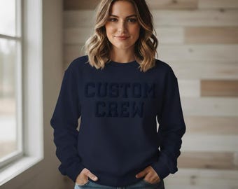 May include: A navy blue crewneck sweatshirt featuring the text CUSTOM CREW embossed in a matching dark blue tone across the chest. The garment has long sleeves, a ribbed neckline, and a standard fit, paired with blue denim jeans.