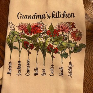 May include: A beige kitchen towel with the text "Grandma's kitchen" in black script. The towel features a floral design with names below the flowers. The names include Marcia, Jackson, Cameron, Kate, Levi, Carter, Noah, and Addilyn.