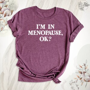 May include: A maroon t-shirt with the text "I'm in Menopause, OK?" printed in white.