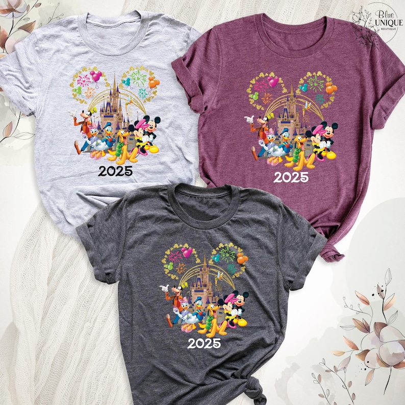 Puede incluir: Tres camisetas con un estampado gr&aacute;fico de Mickey Mouse y sus amigos frente al Castillo de Cenicienta con fuegos artificiales. El texto "2025" est&aacute; impreso en cada camiseta. Las camisetas son de color gris, morado y gris oscuro.