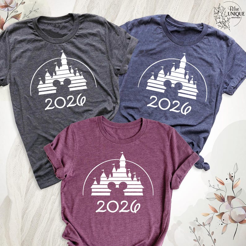 May include: Three short-sleeved t-shirts in charcoal, navy, and burgundy. Each shirt features a white graphic of a castle with Mickey Mouse ears and the year "2026" printed below. The shirts are made of a soft material.