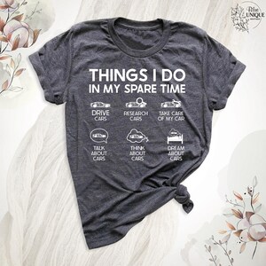 Op de afbeelding: Een donkergrijs t-shirt met de tekst "THINGS I DO IN MY SPARE TIME" en een lijst met autogerelateerde activiteiten, waaronder "DRIVE CARS", "RESEARCH CARS", "TAKE CARE OF MY CAR", "TALK ABOUT CARS", "THINK ABOUT CARS" en "DREAM ABOUT CARS".