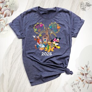 Puede incluir: Una camiseta azul oscuro con un gr&aacute;fico de Mickey Mouse, Minnie Mouse, Donald Duck, Goofy y Pluto frente al Castillo de Cenicienta con fuegos artificiales. El texto "2025" est&aacute; debajo de los personajes.