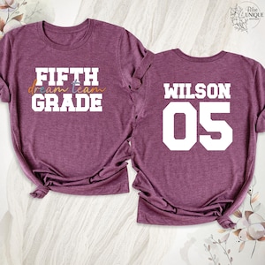 以下が含まれることがあります： 2枚のマルーン色のTシャツ。1枚は白いブロック体で「FIFTH GRADE」、カラフルな筆記体で「dream team」と書かれています。もう1枚は背面に白いブロック体で「WILSON 05」と表示されています。学校に最適です。