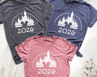 May include: Three short-sleeve crew neck t-shirts in dark gray, navy blue, and mauve. Each shirt features a white graphic of a castle silhouette with a mouse ear icon at the base and the text 2026 printed underneath the design.