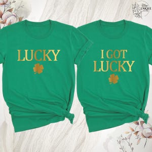 K&ouml;nnte beinhalten: Zwei gr&uuml;ne T-Shirts mit goldenen Buchstaben und Kleeblatt-Grafiken. Ein Shirt sagt "LUCKY" und das andere sagt "I GOT LUCKY". Die Shirts sind aus weichem Material und ideal f&uuml;r St. Patrick's Day.