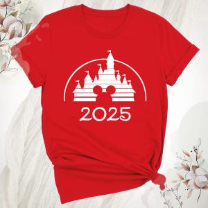 May include: A red t-shirt with a white graphic of a castle, Mickey Mouse ears, and the year 2025. The design is set within a white semi-circle. The shirt is made of a soft material.