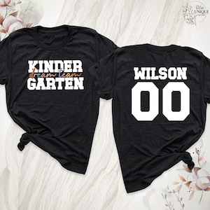 以下が含まれることがあります： 2つの黒いTシャツ。1つは、白、オレンジ、青、紫で「KINDER dream team GARTEN」というテキストが特徴です。もう1つは、白で「WILSON」と数字「00」を表示しています。どちらのシャツも裾が結ばれています。