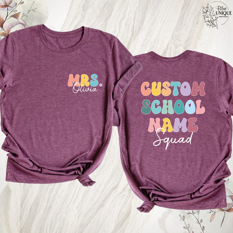 May include: Two maroon t-shirts with rolled sleeves. One shirt has "MRS. Olivia" in colorful text. The other shirt has "CUSTOM SCHOOL NAME Squad" in colorful text. The shirts are on a white surface.
