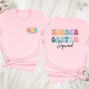 May include: Two light pink t-shirts. One shirt has "MRS. Olivia" in colorful letters on the front. The other shirt has "KINDERGARTEN Squad" in colorful letters on the back. The shirts are likely for a teacher and her class.