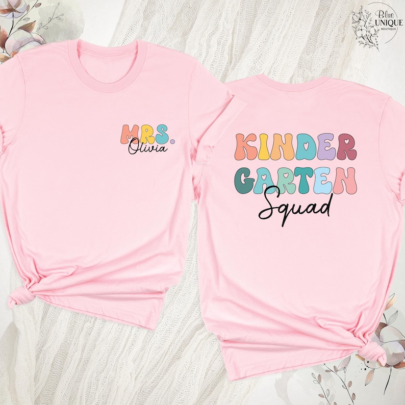 May include: Two light pink t-shirts. One shirt has "MRS. Olivia" in colorful letters on the front. The other shirt has "KINDERGARTEN Squad" in colorful letters on the back. The shirts are likely for a teacher and her class.
