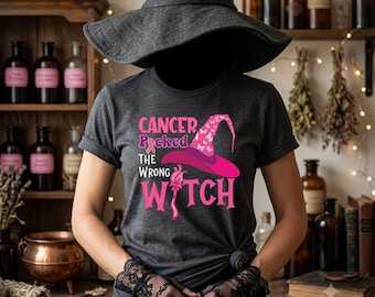 May include: A dark gray short-sleeve t-shirt featuring a pink graphic of a witch hat and the text Cancer Picked The Wrong Witch. The shirt is worn with black lace fingerless gloves and a wide-brimmed black witch hat.