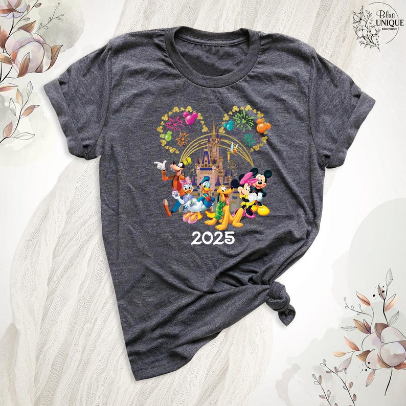 Puede incluir: Una camiseta gris oscuro con un gr&aacute;fico de Mickey Mouse, Minnie Mouse, Donald Duck, Goofy, Pluto y Daisy Duck frente al Castillo de Cenicienta con fuegos artificiales en forma de orejas de Mickey Mouse. El texto "2025" est&aacute; impreso debajo del gr&aacute;fico.