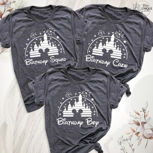 Pode incluir: Tr&ecirc;s t-shirts cinzas com uma silhueta branca do castelo da Cinderela e fogos de artif&iacute;cio. As t-shirts dizem "Birthday Squad", "Birthday Crew" e "Birthday Boy".