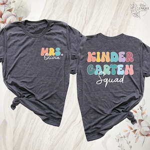 May include: Two charcoal gray t-shirts with colorful text. One shirt says "MRS. Olivia" in pastel colors. The other says "KINDER GARDEN Squad" in a similar font. Both shirts are knotted at the bottom.