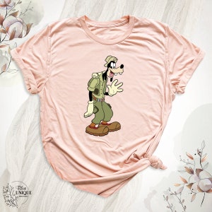 May include: A pink t-shirt with a cartoon image of Goofy dressed as a park ranger. Goofy is wearing a green shirt, brown pants, a brown hat, and a brown backpack. He is waving with his right hand.