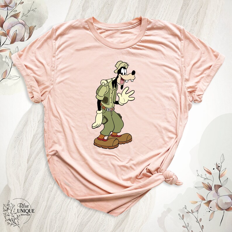 May include: A pink t-shirt with a cartoon image of Goofy dressed as a park ranger. Goofy is wearing a green shirt, brown pants, a brown hat, and a brown backpack. He is waving with his right hand.