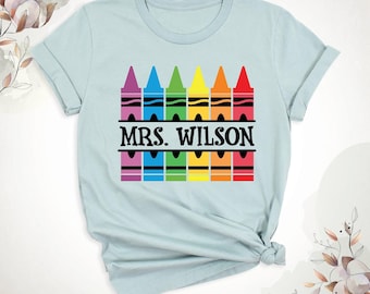 May include: A light blue short-sleeve t-shirt featuring a graphic of six colorful crayons arranged in a row. The text MRS. WILSON is printed in black capital letters across the center of the crayons, which are purple, blue, green, yellow, orange, and red.