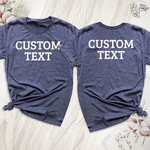 Może przedstawiać: Dwie granatowe koszulki z białym napisem "CUSTOM TEXT" z przodu i z tyłu.