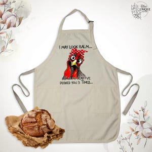Puede incluir: Delantal de cocina beige con un bolsillo. El delantal presenta una ilustraci&oacute;n de dibujos animados de una gallina con una bandana roja y el texto "Puedo parecer tranquilo... Pero en mi cabeza te he picado 3 veces..."