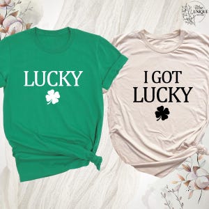K&ouml;nnte beinhalten: Zwei T-Shirts, eines gr&uuml;n und eines beige, mit Text zum Thema St. Patrick's Day und Kleeblatt-Grafiken. Das gr&uuml;ne Shirt sagt "LUCKY" mit einem wei&szlig;en Kleeblatt. Das beige Shirt sagt "I GOT LUCKY" mit einem schwarzen Kleeblatt.