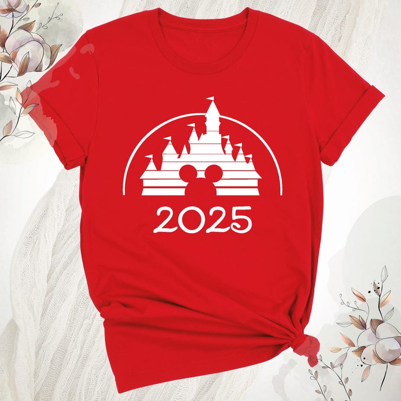 May include: A red t-shirt with a white graphic of a castle, Mickey Mouse ears, and the year 2025. The design is set within a white semi-circle. The shirt is made of a soft material.