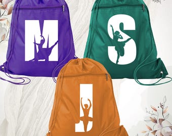 May include: Three drawstring backpacks in purple, teal, and orange. Each bag features a large white letter—M, S, or J—with a silhouette of a dancer integrated into the negative space of the letter. Each bag has a horizontal zippered pocket.