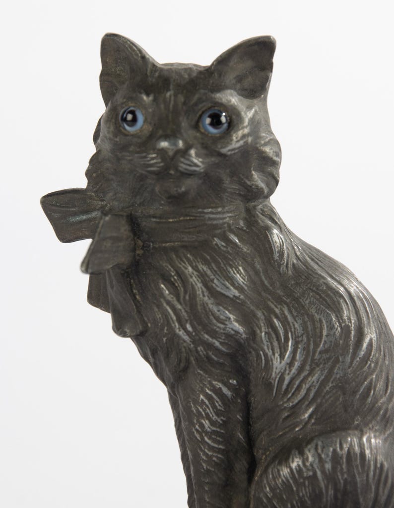 May include: A detailed, antique-style cat figurine with a dark, metallic finish. The cat has bright blue eyes and a bow around its neck. The figurine is a decorative item, likely for display.