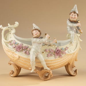 Puede incluir: Una jardinera decorativa de porcelana con forma de barco con dos figuras. El barco es de color crema con detalles dorados, rosas rosas y detalles azules. Las figuras visten trajes y sombreros blancos, sosteniendo objetos dorados.