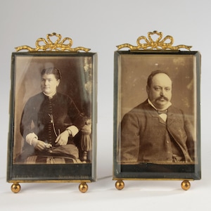 Könnte beinhalten: Zwei antike Porträtrahmen, jeweils mit einer sepiafarbenen Fotografie. Die Rahmen haben oben ein goldfarbenes Bogendesign und kleine goldene Kugelfüße. Ein Rahmen zeigt eine Frau, der andere einen Mann.