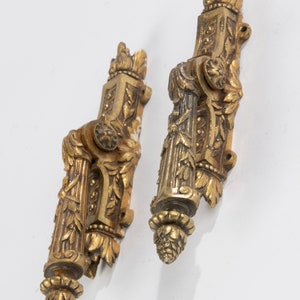 Puede incluir: Dos piezas de herrajes de puerta o ventana de metal dorado ornamentado con intrincados diseños florales. Cada pieza tiene un remate decorativo en la parte superior e inferior.