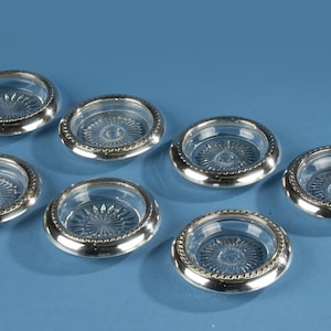 May include: Seven clear glass coasters with silver-colored rims. Each coaster has a starburst design in the center. The coasters are arranged on a blue surface.