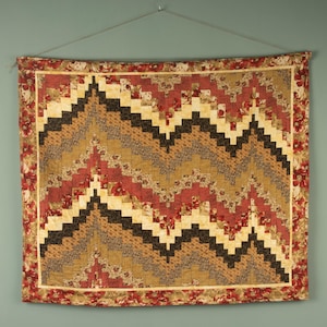 May include: A rectangular wall quilt featuring a chevron pattern composed of small fabric squares in shades of cream, tan, dark brown, and floral red. The quilt is framed by a wide border with a red floral print and hangs from a thin wire.